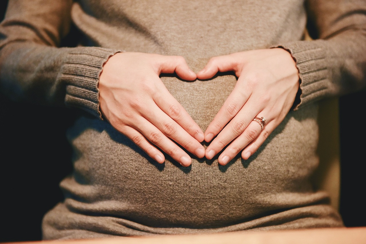 A pregnant woman wearing a brown sweater and forming a heart with her hands over her stomach